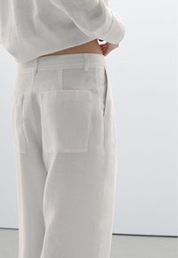 Světle béžové lněné kalhoty s vysokým pasem, rovnými nohavicemi a velkými zadními kapsami. Hladká textura s minimalistickými designovými prvky.