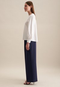 Blusa bianca con maniche a sbuffo, abbinata a pantaloni blu scuro a vita alta. L'outfit presenta un tessuto morbido e una struttura sagomata.