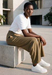 Persoană purtând tricou alb, pantaloni evazați de culoare măslinie și adidași albi, stând pe un bloc de beton în aer liber lângă o clădire modernă.