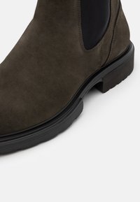 Bottine en suede vert foncé avec des panneaux élastiques latéraux, semelle en caoutchouc noir texturé et accents en cuir lisse sur le talon.