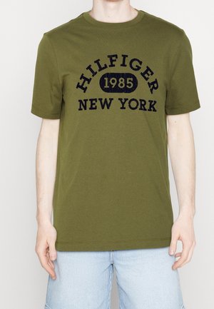 Oliven grøn t-shirt med korte ærmer lavet af bomuld og med rund hals. Har et mørkeblåt logo-tryk: "HILFIGER 1985 NEW YORK."
