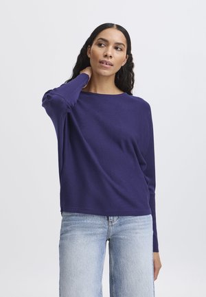 Maglietta lunga viola con un fit rilassato e scollatura a girocollo, realizzata in tessuto morbido. Indossata con jeans denim azzurri.