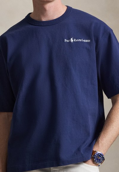 Άνδρας που φορά μπλε μπλουζάκι Polo Ralph Lauren και ρολόι με μπλε καντράν και καφέ δερμάτινο λουράκι στον αριστερό καρπό.