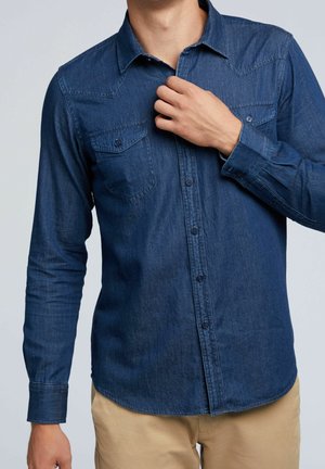 Mann in langärmligem blauen Jeanshemd und beigen Hosen richtet seinen Kragen, Brusttaschen sichtbar, Kopf nicht zu sehen.