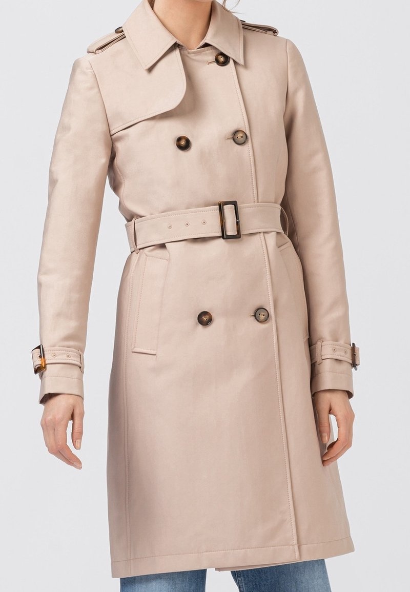 Trench-coat beige à double boutonnage avec ceinture, épaulettes et poignets boutonnés, porté sur un jean bleu.