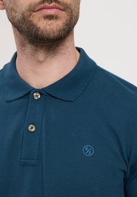 Granatowa koszulka polo wykonana z fakturowanej tkaniny, z klasycznym kołnierzykiem, dwoma złotymi guzikami i małym niebieskim logo na piersi.