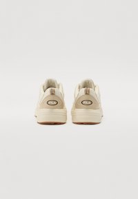 Zapatillas deportivas beige con paneles de malla y talón reforzado. Presentan el logo de Fila en la parte posterior, suelas texturizadas y sutiles acentos marrones.