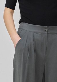 Pantalons plissés gris avec une fermeture à bouton et des poches latérales, associés à un haut noir ajusté. Le tissu a une texture lisse.