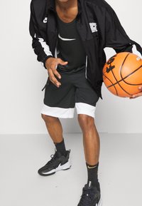 Man wearing black and white Nike sportswear dribbling an orange basketball indoors on a plain background.