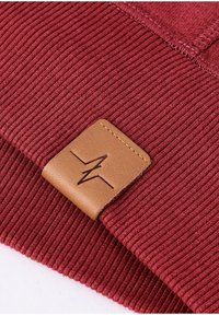 Dieprode geribbelde stof met een tan leren patch met een ingeslagen logo. Gedetailleerd stikwerk zichtbaar aan de randen en op de oppervlaktetextuur.