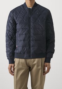 Man som bär en marinblå vadderad bomberjacka med ribbade muddar och fåll, i kombination med beiga byxor, stående mot en enhetlig bakgrund.