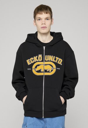 Młody mężczyzna w czarnej bluzie z kapturem na zamek, z żółtym logo "ECKŌ UNLTD." i grafiką nosorożca, ręce w kieszeniach, niebieskie dżinsy, neutralne tło.
