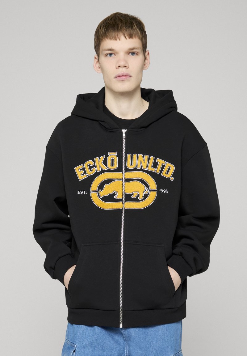 Jeune homme portant un sweat à capuche noir zippé avec le logo jaune "ECKŌ UNLTD." et un dessin de rhinocéros, mains dans les poches, jean bleu, fond neutre.