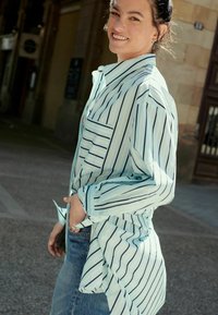 Lyseblå stribet skjorte med sorte og hvide striber, knaplukning foran, enkelt brystlomme, parret med denim jeans. Blødt stof, afslappet pasform.