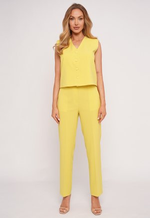 Woman standing wearing sleeveless yellow buttoned top and matching high-waist straight-leg pants with nude open-toe heels.