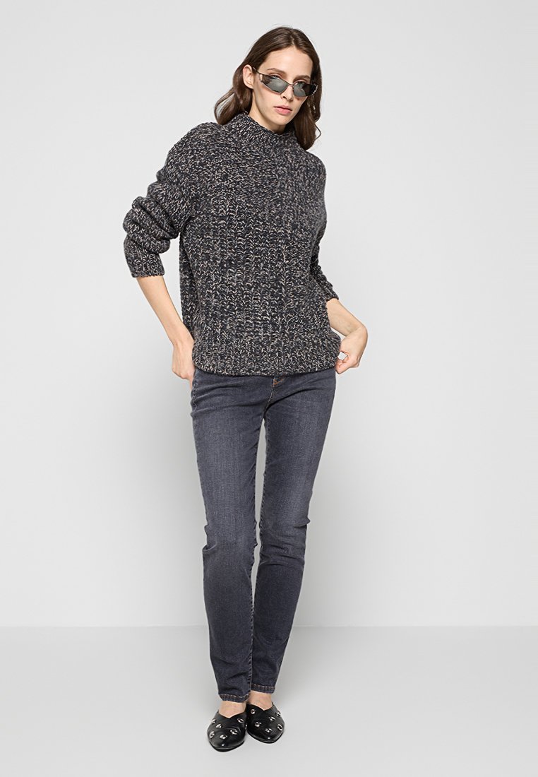 Grauer gestrickter Pullover mit hohem Kragen, kombiniert mit dunklen Jeans und schwarzen Slippern mit Hardware-Akzenten. Das Model posiert vor einem neutralen Hintergrund.