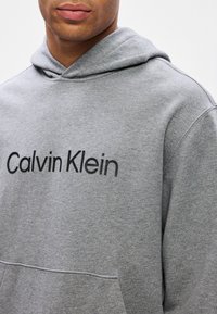 Muž v šedé mikině s kapucí a černým nápisem "Calvin Klein" přes hruď, zastřižené na úrovni úst a ramen.