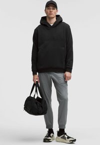 Schwarzer Kapuzenpullover mit geprägtem Logo, graue tapered Hosen, schwarze Sportschuhe mit weißen Akzenten und eine schwarze Reisetasche.