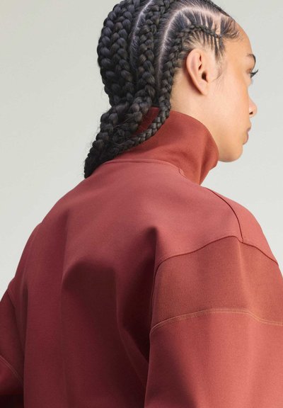 Czerwona bluza z wysokim kołnierzem wykonana z teksturowanej tkaniny, z wyraźnymi przeszyciami i luźnym krojem. Widok od tyłu, z warkoczami.