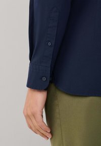 Camisa azul marino abotonada con textura suave, que presenta un cuello puntiagudo, puño abotonado y manga remangada, combinada con pantalones verde oliva.