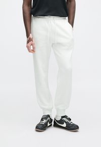 Šviesiai pilkos laisvalaikio kelnės su rankogaliais ties čiurnomis ir raišteliais ties juosmeniu, derinamos su juodais Nike sportbačiais, kuriuose yra baltų akcentų ir tekstūruotas dizainas.