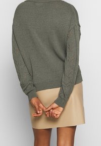 Pull vert olive en tricot avec un ourlet côtelé, présentant des motifs complexes en dentelle sur les manches. Associé à une jupe beige.