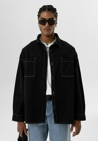 Zwarte denim overhemd met twee voorzakken en witte stiksels, gedragen over een wit t-shirt en gecombineerd met lichtblauwe jeans.