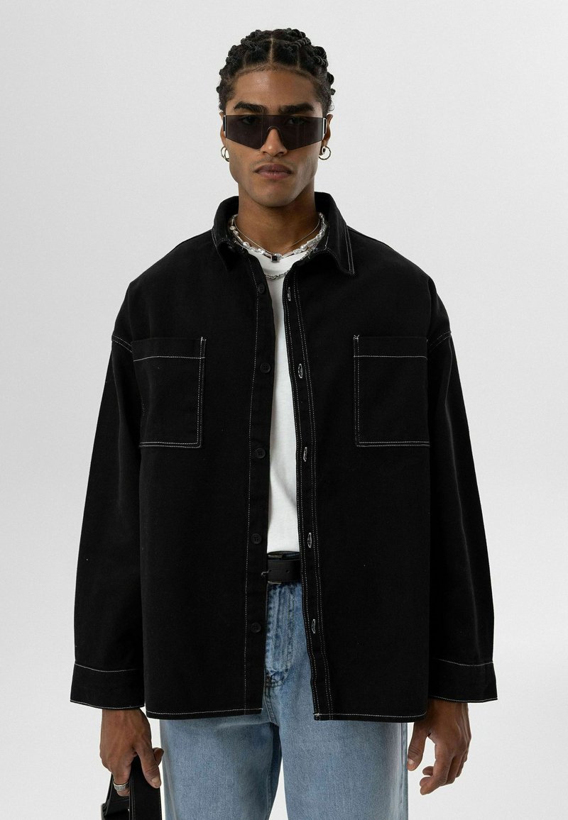 Zwarte denim overhemd met twee voorzakken en witte stiksels, gedragen over een wit t-shirt en gecombineerd met lichtblauwe jeans.