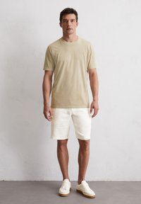 Beige Baumwoll-T-Shirt, kurze Ärmel, Rundhalsausschnitt; kombiniert mit weißen Leinen-Shorts. Das Modell trägt weiße Sneaker mit beigen Akzenten.