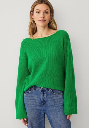 Vrouw draagt een felgroene geribbelde gebreide trui met lange mouwen en blauwe spijkerbroek, staand tegen een lichtgrijze muur.