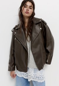 Junge Frau mit langen braunen Haaren, die eine übergroße dunkelbraune Lederjacke über einem grauen Pullover mit Spitzenrand und blauen Jeans trägt.