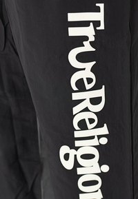Schwarze Hose mit einem großen weißen "True Religion"-Logo, das vertikal an der Seite verläuft und eine glatte, matte Oberfläche zeigt.