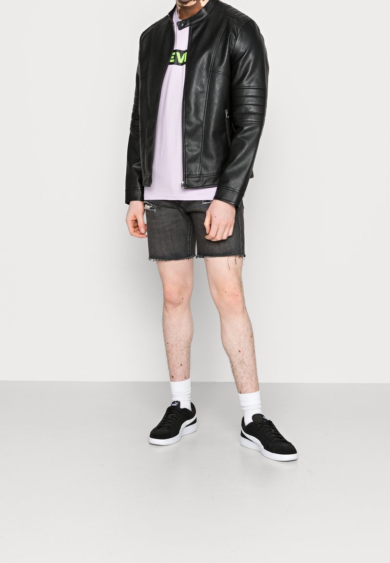 Personne portant une veste en cuir noire, un t-shirt violet clair, un short en jean noir déchiré, des chaussettes blanches et des baskets noires, debout contre un mur uni.