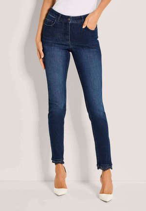 Frau trägt dunkelblaue Skinny-Jeans mit ausgefransten Säumen und weiße spitze Pumps, steht vor einem neutralen Hintergrund.