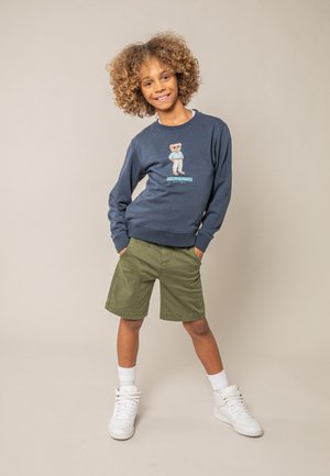 Enfant aux cheveux bouclés portant un sweat-shirt bleu marine avec un motif d'ours en peluche, un short olive, des chaussettes blanches et des baskets blanches, debout les mains dans les poches.