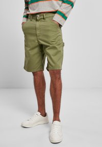 Man med olivgröna cargoshorts, randig långärmad skjorta och vita sneakers, med händerna i fickorna, stående på en grå golv.