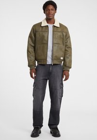 Giacca bomber verde oliva con colletto in ecopelliccia color crema, chiusura con zip e due tasche laterali, abbinata a pantaloni cargo grigi e stivali neri.