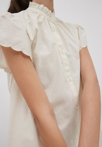 Cremfarbendes Blusenshirt mit kurzen Puffärmeln, gerüschtem Kragen und Knopfverschluss. Glattes Material mit gewellten Ärmelabschlüssen.