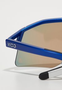 Lunettes de soleil bleues avec une monture géométrique, des verres miroités, et la marque "KCO" sur le côté. Présente un contraste entre textures mates et brillantes.
