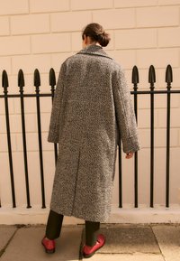 Manteau long en pied-de-poule gris et noir avec un col oversized, posé contre un mur clair et une balustrade en fer forgé noir. Des chaussures rouges sont visibles.