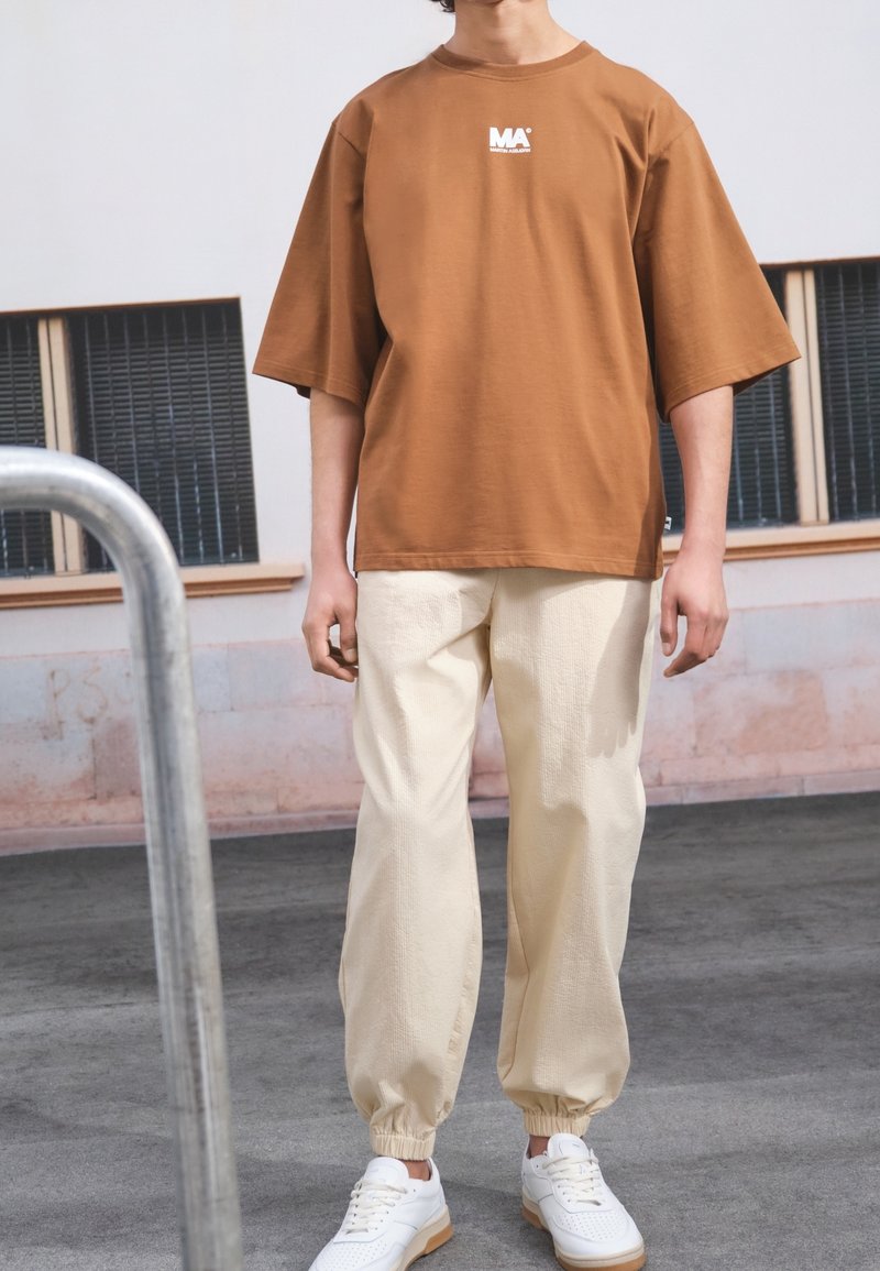 Camiseta marrón de gran tamaño con un logo blanco, combinada con pantalones elásticos de color beige claro con puños, y zapatillas blancas con acentos beige.