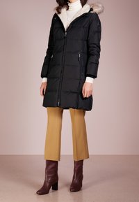 Svart puffjacka med en furfoder på huvan, med dragkedja fram, sidofickor och en åtsittande silhuett, i kombination med beige byxor och bruna stövlar.