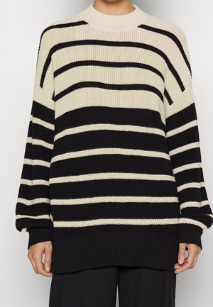 Personne portant un pull en maille rayé noir et blanc à manches longues avec un ourlet côtelé, associé à un pantalon noir.