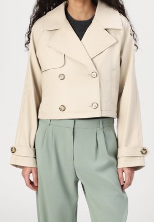 Woman wearing beige cropped double-breasted jacket with wide lapels and buttoned cuffs, paired with high-waisted light green pleated trousers.