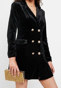 Czarna aksamitna sukienka-marynarka o kroju dwurzędowym, z złotymi guzikami i przednimi kieszeniami. W zestawie z kopertówką ze złotym brokatem.