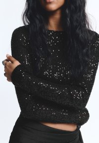 Fekete flitteres pulóver hosszú ujjakkal, texturált kötött anyagból és testhez simuló kialakítással. Párban egy fekete szoknyával, amely a letisztult stílust hangsúlyozza.