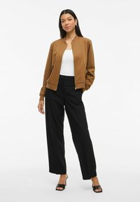 Femme debout dans une veste zippée marron, un haut blanc, un pantalon large noir et des sandales à talons noires contre un fond blanc uni.