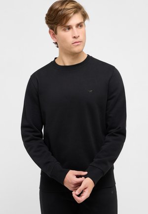 Junger Mann mit hellbraunen Haaren, trägt ein schwarzes Sweatshirt mit langen Ärmeln, schaut nach links, die Hände vor sich verschränkt.