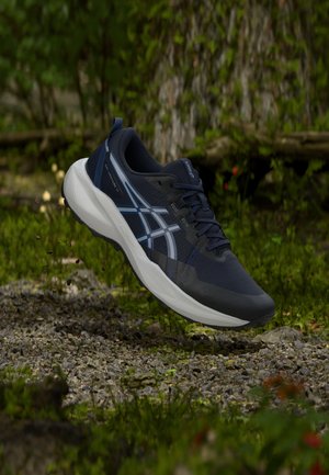 Chaussure de course bleu foncé avec semelle blanche et bandes, capturée en plein vol au-dessus d'un chemin de gravier dans un cadre forestier verdoyant.