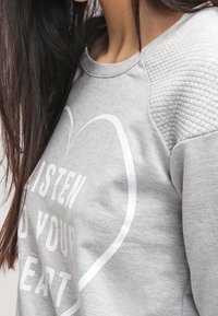 Donna con capelli lunghi e scuri che indossa un felpa grigia chiara con maniche testurizzate e un contorno a forma di cuore bianco attorno al testo "Ascolta il tuo cuore."